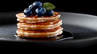 Stack of golden mini pancakes topped with fresh blueberries and mint leaf on dark plate against black background for breakfast menu design.