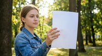 Young woman in denim jacket reading document outdoors in sunny park setting with green trees background for business and lifestyle concepts.