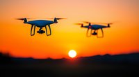 Flying drones silhouetted against vibrant orange sunset sky with glowing sun on horizon, modern quadcopters in dramatic aerial photography scene.