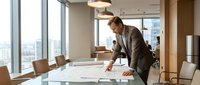 Professional businessman in suit analyzing architectural plans and documents on conference table in modern office with city skyline view through windows.