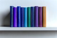 Row of colorful hardcover books standing on white shelf against light background, arranged in blue, purple and orange tones creating minimalist composition.