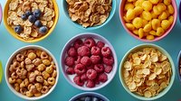 Colorful bowls filled with breakfast cereals, fresh raspberries, blueberries and crunchy snacks on turquoise background arranged in neat rows.