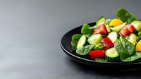 Fresh summer salad with spinach leaves, sliced cucumber, cherry tomatoes and yellow bell pepper on black plate against gray background. Food photography.