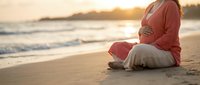 Pregnant woman in coral sweater sitting peacefully on sandy beach at golden sunset, expecting mother enjoying serene coastal moment during maternity.