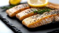 Grilled salmon fillets seasoned with herbs and black pepper served on dark slate board with fresh rosemary sprigs and lemon slices, close up view of cooked fish.