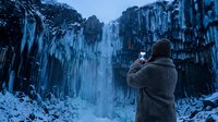 Traveler in winter coat photographing frozen waterfall with columnar basalt formations, capturing nature's icy landscape during cold season adventure.