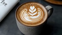 Creamy latte art featuring delicate leaf pattern in frothy milk foam, served in modern gray ceramic cup against dark background. Perfect for cafe marketing.