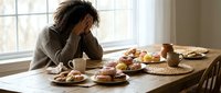 Young African American woman feeling overwhelmed during breakfast at wooden dining table with healthy meal and coffee cup in bright kitchen.