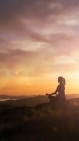 Woman meditating in lotus pose on mountain peak during golden hour sunset with dramatic pink and orange sky creating peaceful spiritual atmosphere.