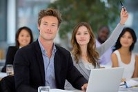 Young Caucasian business professionals in modern office meeting with diverse colleagues, woman raising hand to participate in discussion.