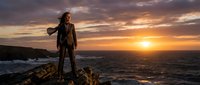 Young adventurous woman hiker standing on rocky cliff overlooking ocean at golden sunset. Outdoor adventure and travel lifestyle concept for active tourism.