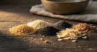 Assorted ancient grains and seeds scattered on rustic wooden surface with ceramic bowl and linen cloth creating warm kitchen atmosphere for healthy cooking.