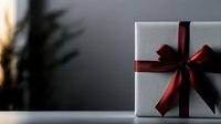 White gift box with elegant burgundy red satin ribbon and bow on dark background with soft focus plant silhouette, festive holiday presentation.