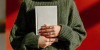 Person in green knit sweater holding white book or journal, with manicured nails and gold rings, against red background.