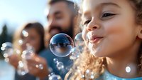 Young girl with curly hair blowing soap bubbles outdoors on sunny day, close-up portrait with family in background, capturing joyful childhood moment.