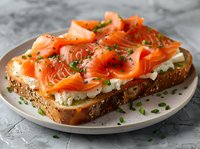 Fresh salmon slices layered on whole grain toast with cream cheese spread, garnished with chopped chives and black pepper on white ceramic plate.