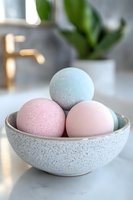 Pastel bath bombs in speckled ceramic bowl on marble counter, soft natural lighting creates gentle shadows. Mint and pink colors with plant decor in background.