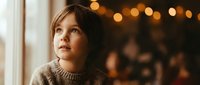 Young Caucasian boy in cozy sweater gazing thoughtfully by window with warm bokeh lights creating magical holiday atmosphere for family moments.