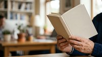 Senior adult with light skin tone reading open book indoors, home library setting with bookshelves and plants visible, leisure and education concept.