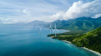 Offshore wind turbines along tropical coastline with mountains and blue ocean waters, aerial view showing renewable energy generation and sustainable power.