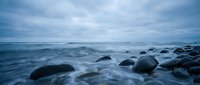 Serene ocean waves washing over smooth dark rocks on coastline during moody blue hour with dramatic cloudy sky creating peaceful seascape atmosphere.