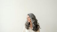 Mature woman with long gray wavy hair and fair complexion, wearing white blazer, smiling while looking to the side against neutral background for professional portraits.