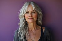 Confident mature woman with silver gray wavy hair smiling against purple background, wearing black top and delicate necklace, radiating natural beauty and wisdom.