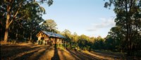 Rustic wooden farmhouse with extended veranda nestled among tall eucalyptus trees during golden hour, casting long shadows across the rural property landscape.