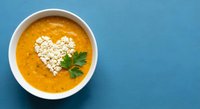 Creamy orange soup with heart-shaped croutons and fresh parsley garnish in white bowl on blue background for healthy comfort food recipes.