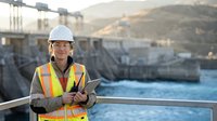 Professional female engineer in safety gear holding tablet at hydroelectric dam construction site during golden hour sunset with mountain landscape.