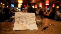 Handwritten apology note on a napkin at a bar counter with a candle and pen, blurred restaurant background with warm bokeh lights.