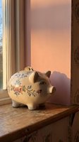 Vintage ceramic piggy bank with floral pattern sits on wooden windowsill in warm golden sunlight creating cozy home savings and financial planning atmosphere.