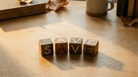 Wooden letter blocks spelling SAVE on hardwood floor with warm lighting and autumn leaves creating cozy financial planning atmosphere for budgeting concepts.