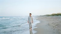 Man in casual linen clothing walking along sandy beach shore with ocean waves, suitable for lifestyle, travel, and wellness marketing campaigns.