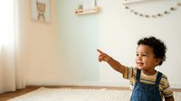 Young child with dark curly hair and medium skin tone wearing denim overalls and striped shirt, pointing and smiling in bright nursery room with natural light, toys and decorations for childcare and parenting content.
