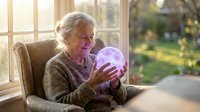 Senior Caucasian woman sitting by window holding glowing crystal ball with mystical purple light in cozy living room setting for fortune telling concept.