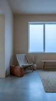 Minimalist interior space featuring a gray fabric lounge chair, vintage brown leather suitcase, and potted dried branches beside a large window with natural light flooding the room.