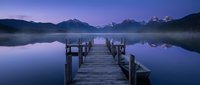 Wooden pier extending into calm mountain lake at twilight with purple sky and snow-capped peaks reflecting in still water creating peaceful landscape scene.