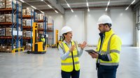 Two warehouse workers in safety vests and hard hats discussing logistics operations in modern distribution center with shelving systems and equipment.