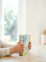 Young child with light skin tone holding colorful stacked books on wooden table near bright window, promoting early literacy and educational development for children.
