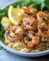 Grilled jumbo shrimp served on orzo pasta garnished with fresh herbs, black pepper and lemon wedges on white plate, close up view of Mediterranean cuisine.