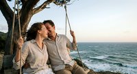 Happy couple enjoying romantic sunset moment on tree swing by ocean shore during golden hour vacation getaway.