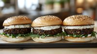 Three juicy beef burgers with creamy sauce and fresh lettuce on sesame buns arranged in row on dark wooden serving board against blurred restaurant background.