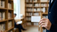 Mature male professional in dark business suit holding blank white card in library setting with bookshelves, suitable for corporate identity and business communication concepts.