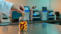 Pharmacist hand reaching for colorful medication capsules in glass jar at modern pharmacy counter with blurred equipment background.