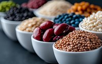 Variety of colorful legumes, beans, lentils and grains in white ceramic bowls arranged in row on dark background. Fresh organic protein sources for healthy eating.