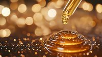 Golden drop of serum falling onto shimmering liquid surface creating ripple effect against warm bokeh background, macro cosmetic photography.