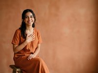 Young woman with long dark hair and medium skin tone, wearing rust-colored linen dress, sitting on wooden stool against terracotta textured wall, smiling with hand on chest, suitable for lifestyle and portrait use.