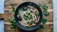 Asian noodle soup with mushrooms and fresh herbs in black bowl on rustic wooden cutting board. Comfort food meal ready to serve.