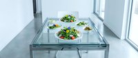 Fresh green salads with edible flowers served on white plates atop a modern glass dining table in a bright minimalist interior.